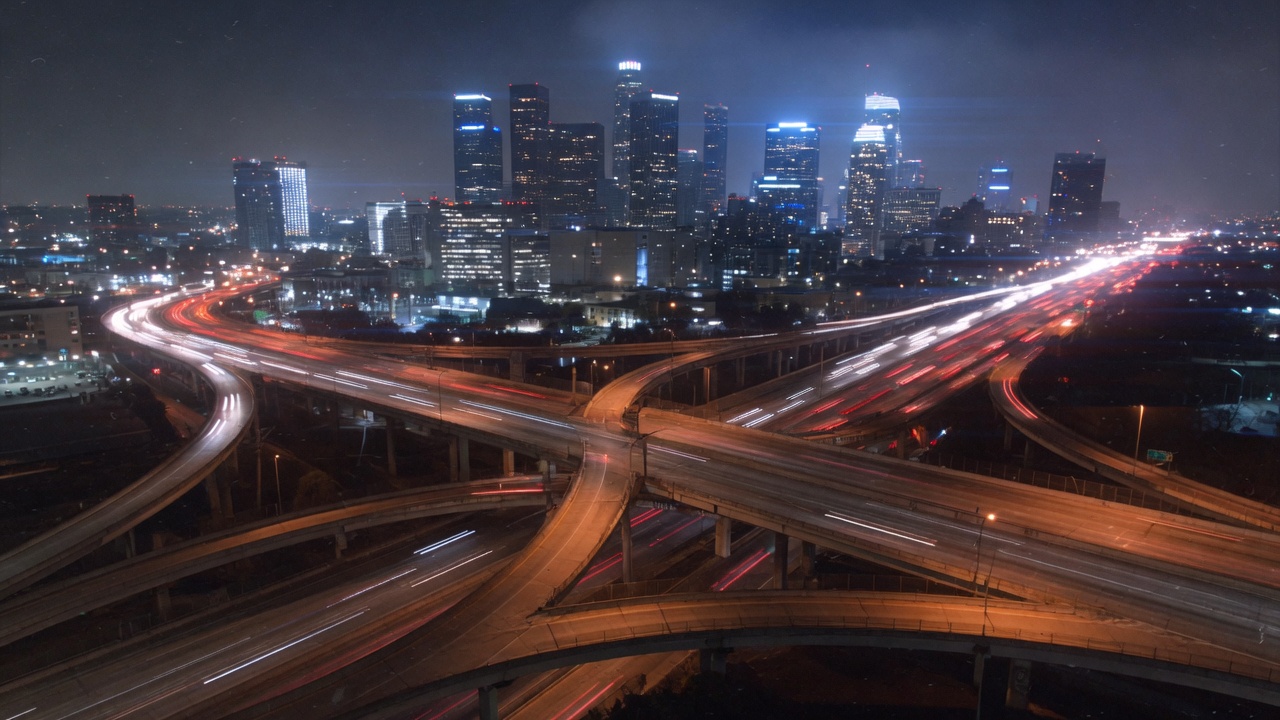 Los Angeles freeway interchange at night, downtown glowing steel-blue in the distance.